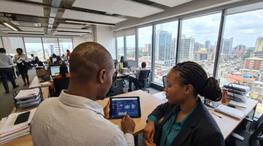 Two professionals in a bright contemporary office space reviewing documents on a tablet, natural interaction captured over-the-shoulder with cityscape visible through large windows